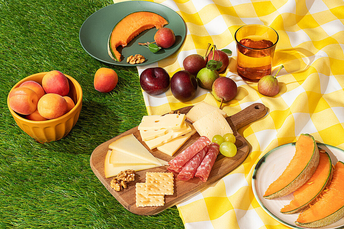 Ein lebhaftes Sommerpicknick mit einer Auswahl an Früchten, Käse und Fleisch, die ordentlich auf einer gelb-weiß karierten Decke auf Kunstrasen angeordnet sind