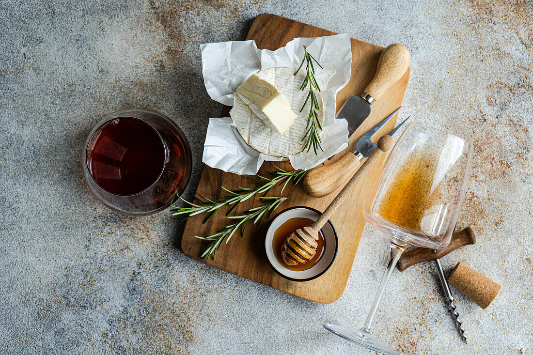 An overhead shot of Brie cheese, fresh rosemary, honey, and a glass of red wine arranged on a rustic wooden board.