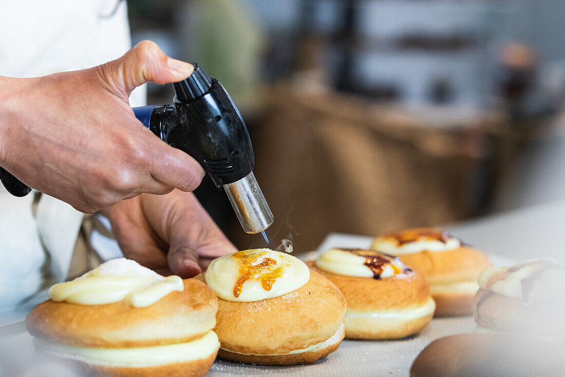 Unrecognizable crop chef caramelizing sugar cream on sweet Berliners with gas torch while preparing vegan dessert in bakehouse