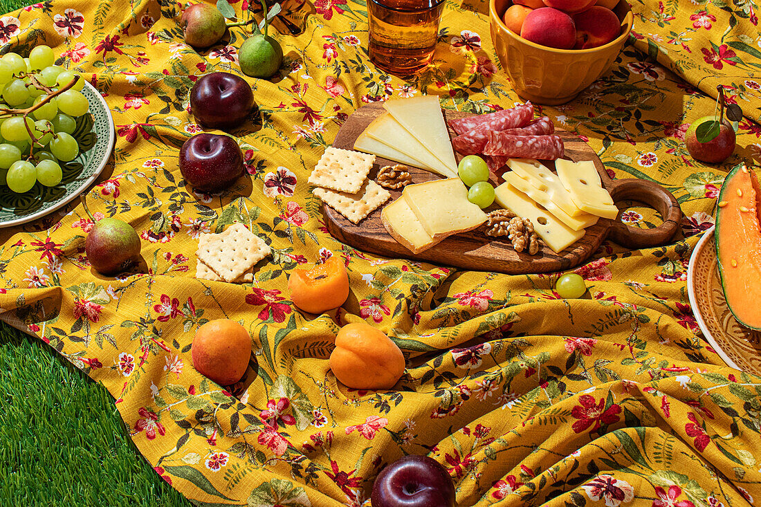 Eine farbenfrohe Auslage mit sommerlichen Picknickgerichten auf einer gelben Blumentischdecke. Das Sortiment umfasst frisches Obst, Käse, Fleisch und Cracker