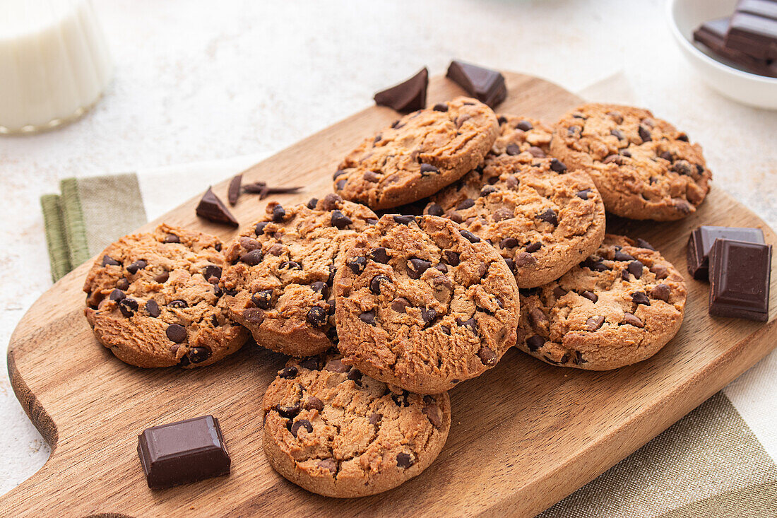 Freshly baked chocolate chip cookies arranged on a wooden cutting board, accompanied by milk and chocolate pieces