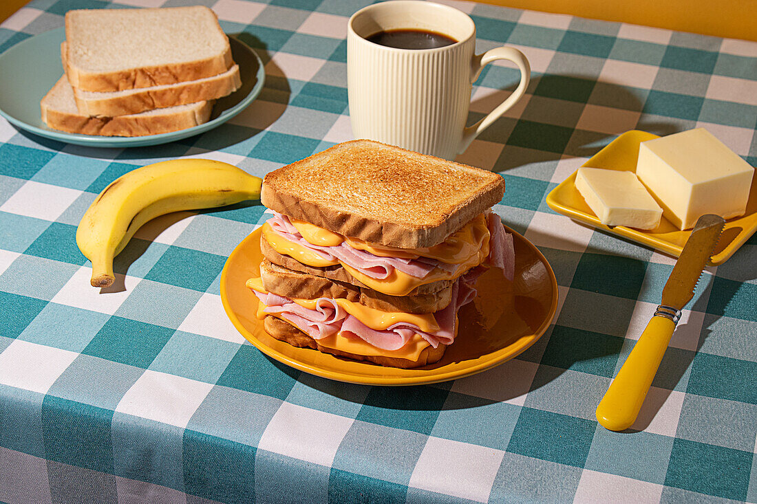 Sandwich auf einem gelben Teller, dazu eine Tasse Kaffee, Butter auf einem kleinen Teller und eine frische Banane, alles auf einer blau-weiß karierten blauen Tischdecke angeordnet