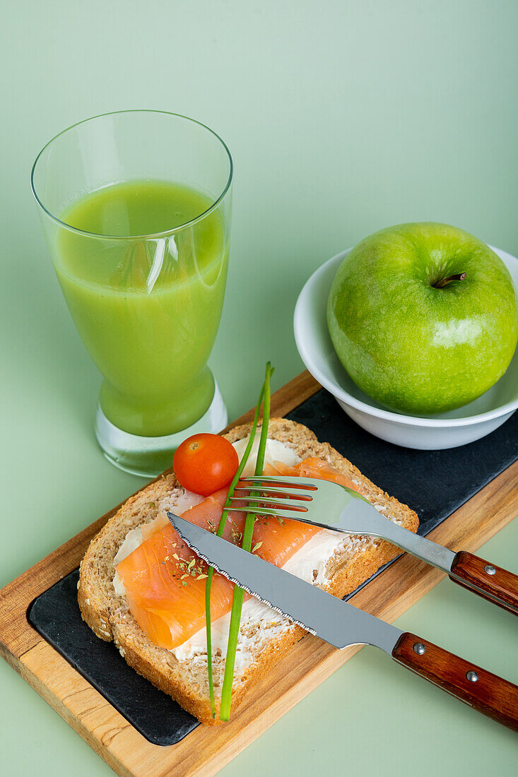 From above nutritious breakfast setup featuring a glass of cucumber and apple juice, a slice of whole grain bread topped with salmon and cream cheese, garnished with a chive and cherry tomato, alongside a fresh green apple.