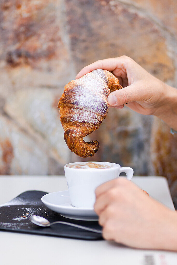 Eine abgeschnittene, nicht erkennbare Person, die ein traditionelles Frühstück mit Kaffee kolumbianischen Ursprungs und einem frisch gebackenen, hausgemachten Croissant mit Puderzucker genießt.