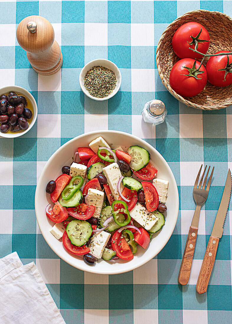 Eine lebendige Schüssel mit griechischem Salat mit Fetakäsewürfeln, geschnittenen Tomaten, Gurken und Oliven auf einem blau-weiß karierten Tischtuch Ein Korb mit Tomaten, eine Schüssel mit Oliven und Gewürzen begleiten diese Szene