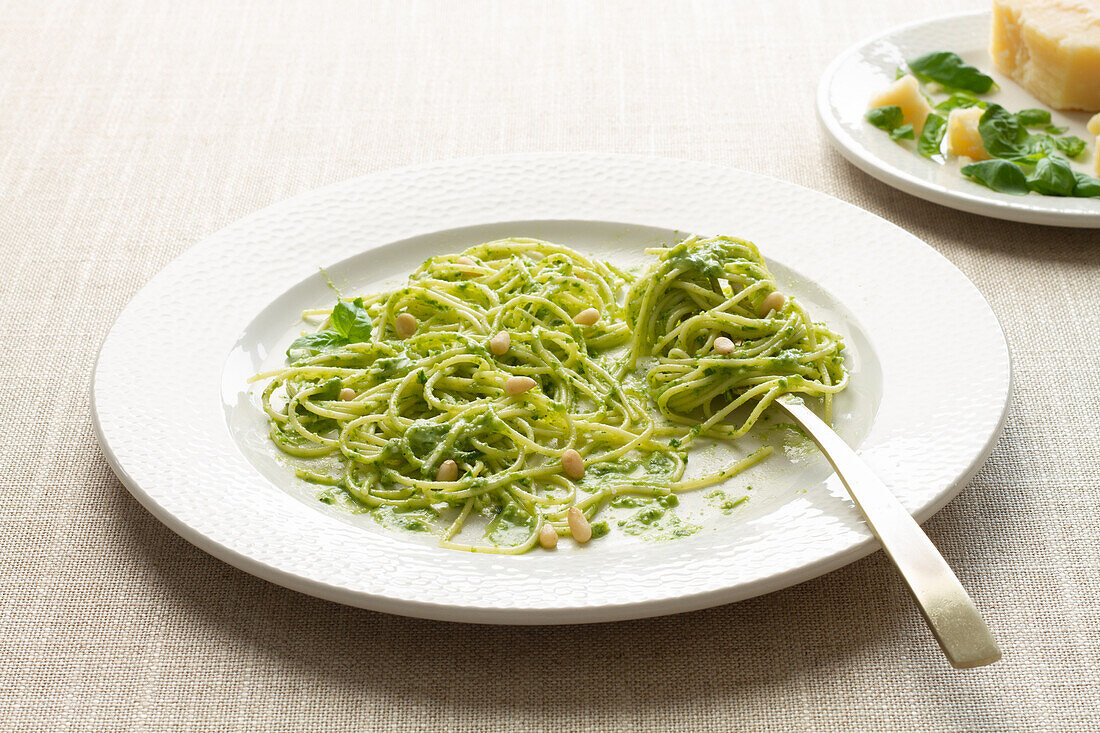 Teller mit Spaghetti in einer reichhaltigen grünen Pestosauce, garniert mit Pinienkernen, auf einem strukturierten Tischtuch, mit Parmesankäse im Hintergrund.