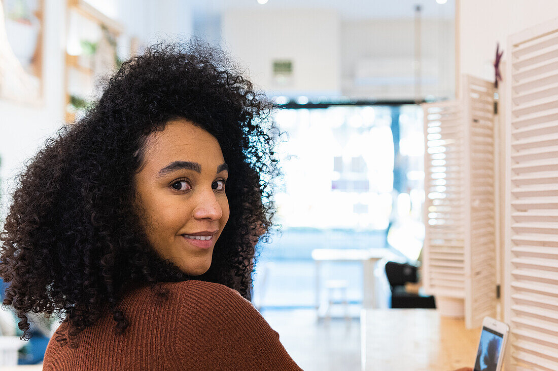 Glückliche Afroamerikanerin mit lockigem Haar, die mit einem Handy am Tresen einer Bar sitzt und über die Schulter in die Kamera schaut.