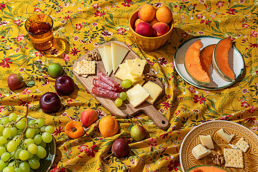 A vibrant spread of fresh fruits, assorted cheeses, salami, nuts, and beverages laid out on a floral-patterned yellow tablecloth, capturing the essence of a summer picnic