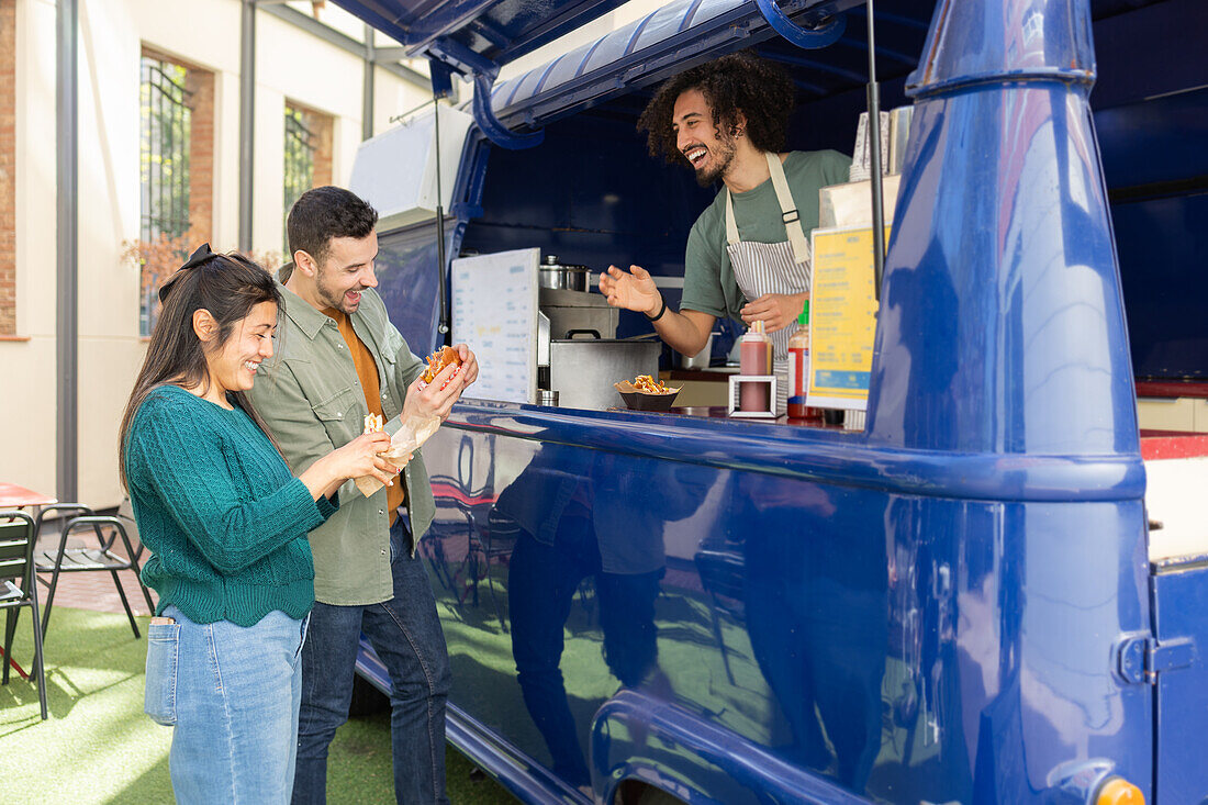 A group of friends share a laugh while purchasing fast food from a cheerful vendor inside a vibrant blue food truck