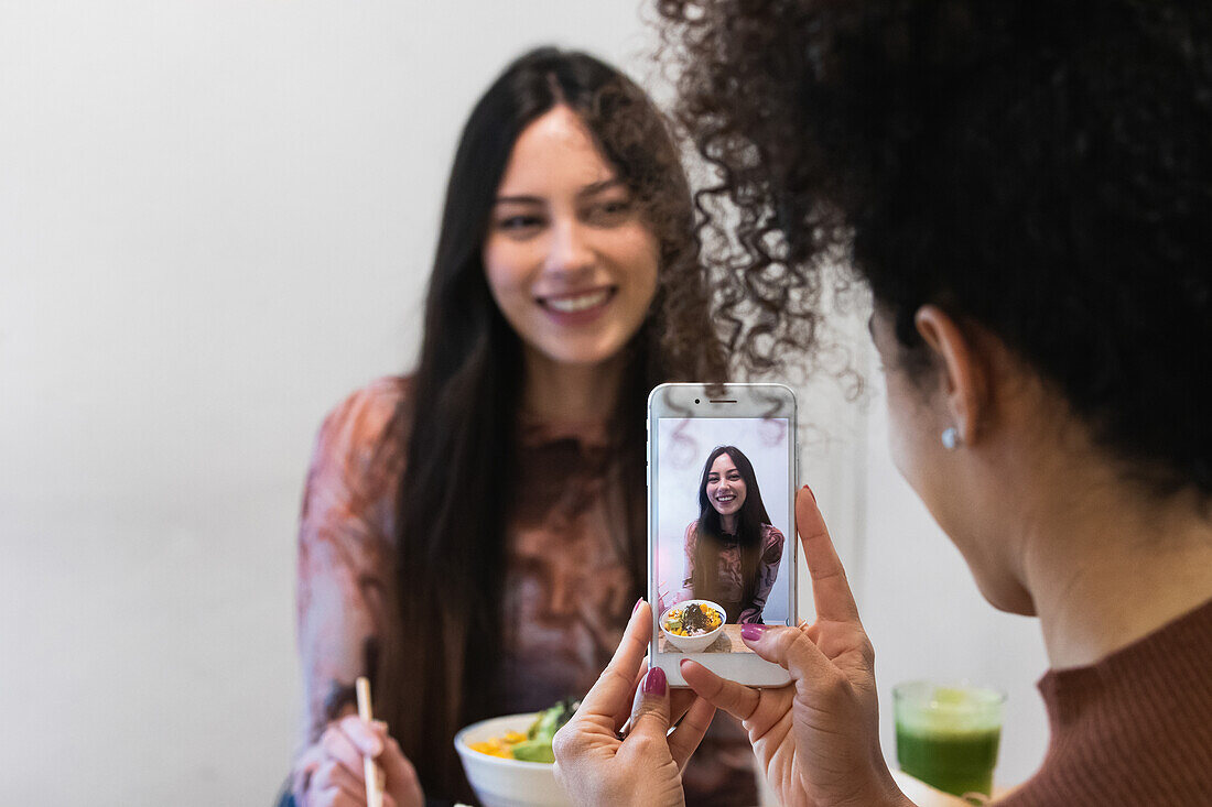 Eine nicht erkennbare Afroamerikanerin fotografiert einen Freund, der mit einem Poke-Gericht in einem Restaurant am Tisch sitzt