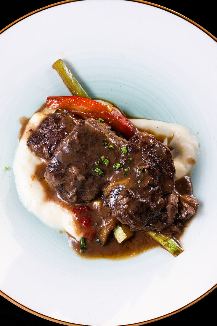 Top view of tantalizing pork cheeks paired with a rich Pedro Ximenez reduction, elegantly presented on a creamy mashed potato base surrounded by sauteed seasonal vegetables.