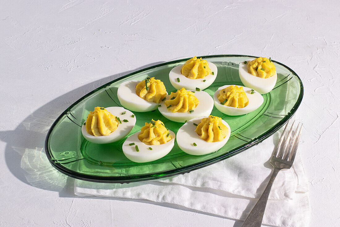 From above a green glass serving plate holding six deviled eggs, garnished with sprinkled chives. The setting includes a white textured background and a silver fork by the side, highlighting a fresh, summery table setting.