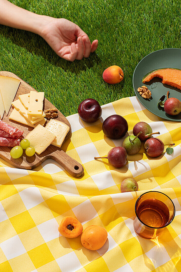 Anonymous person's hand is visible colorful summer picnic scene capturing a variety of fresh fruits like apples, apricots, and plums, alongside assorted cheeses and meats on a wooden board, all set on a yellow and white gingham blanket