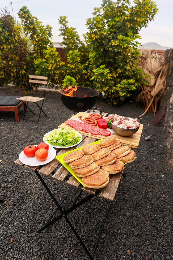 A vibrant setup for an outdoor barbecue featuring a variety of foods including sausages, salads, and buns on a rustic wooden table, with a flaming grill in the background surrounded by lush greenery.