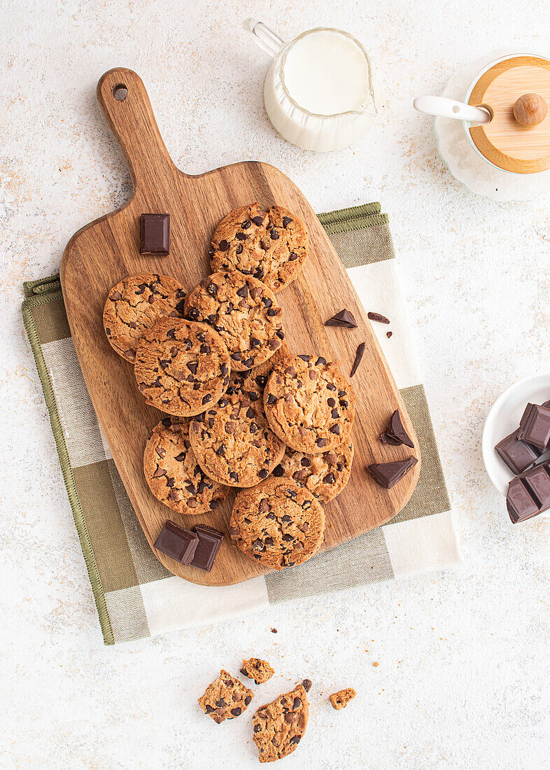 Eine köstliche Auswahl an Schokoladenkeksen auf einem Holzbrett, begleitet von Schokoladenstückchen, einem Milchglas und einer Zuckerdose im Hintergrund - perfekt für Dessert- und Snackkonzepte