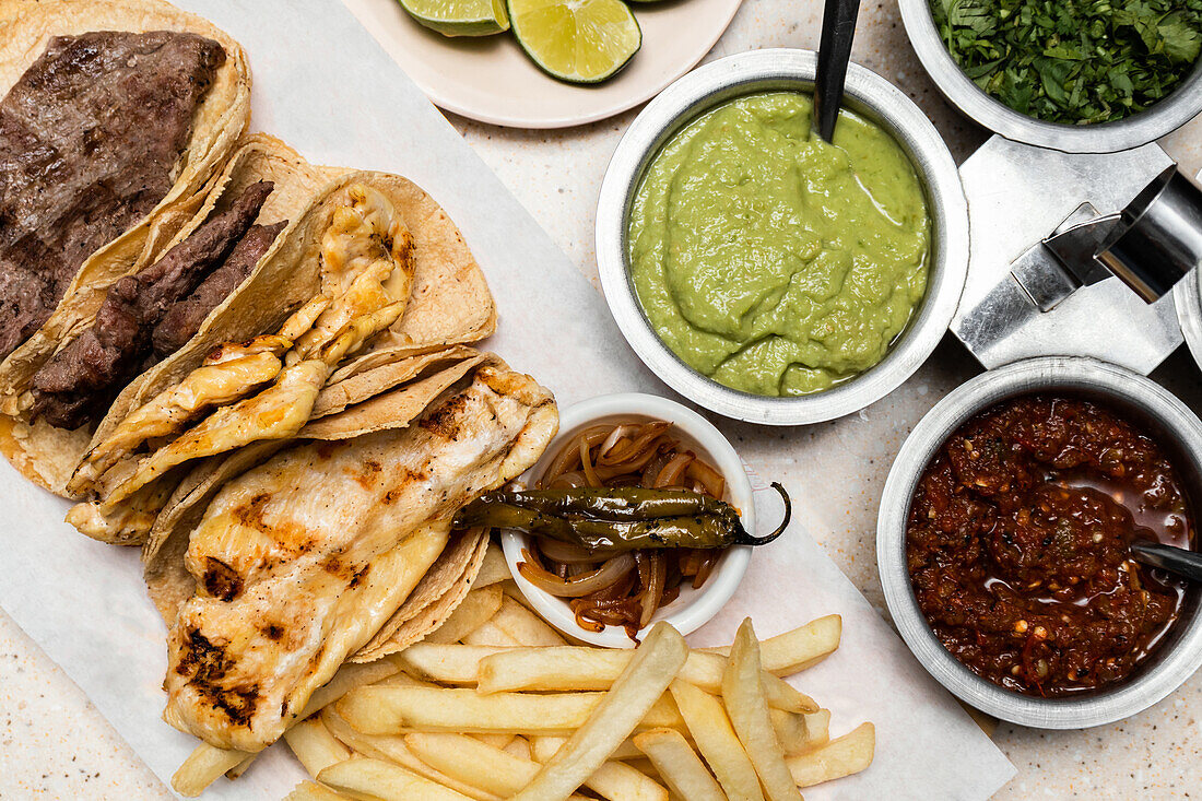 Blick von oben auf ein traditionelles mexikanisches Hausgericht mit gegrilltem Steak auf Maistortillas, dazu Guacamole, Salsa Roja, karamellisierte Zwiebeln und Pommes frites.