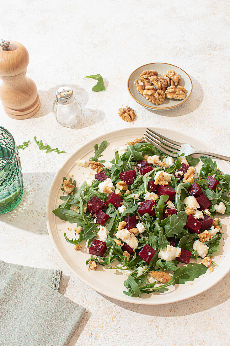 Gesunder Salat aus Ziegenkäse, Rucola und Roter Bete auf einem beigen Teller mit einer Pfeffermühle und einem Kalkwasserglas in einer rustikalen, aber lebhaften Atmosphäre