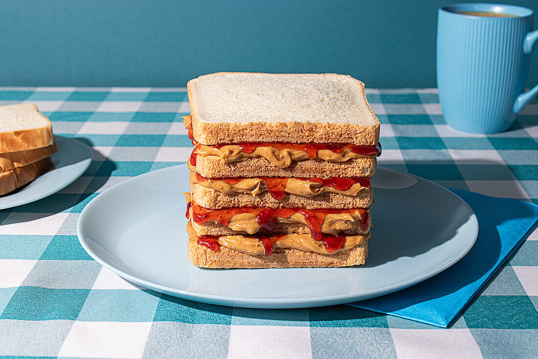 Erdnussbutter-Gelee-Sandwich auf einem blauen Teller, eine Tasse amerikanischer Kaffee und einige Brotscheiben vor einem blauen Hintergrund