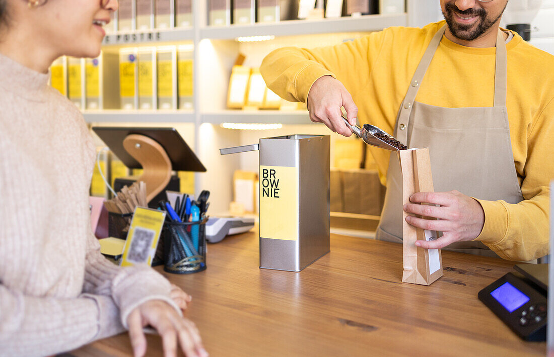 A cheerful barista in a yellow shirt scoops fresh coffee beans for a female customer at a cozy coffee shop.