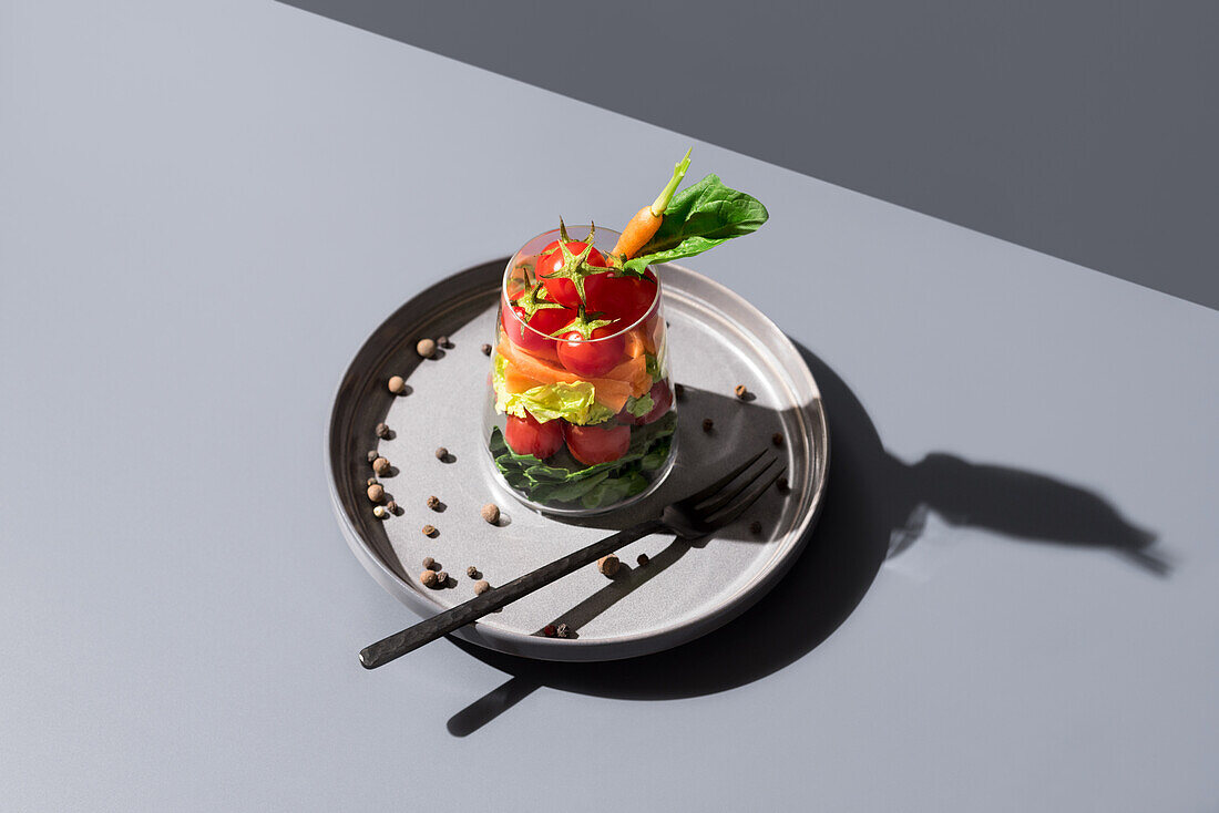 Top view of a layered salad artistically presented in a transparent glass, highlighted by a sharp shadow, on a round metal plate with scattered peppercorns, all set against a grey background.