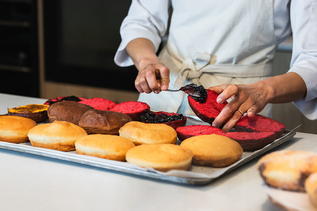 Faceless crop baker spreading sweet jam on vegan sponge cakes while preparing desserts in kitchen of bakery shop