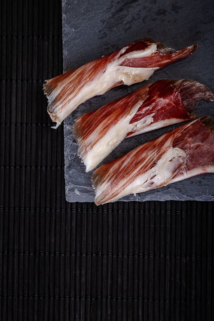 Close-up view of freshly cut serrano ham slices carefully arranged on a slate board, showcasing the marbled texture and rich color contrast on a dark, textured background