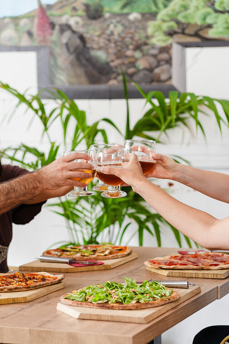 A group of cropped unrecognizable friends enjoying gluten-free beer and gourmet pizzas with a variety of toppings, capturing a moment of joy and good food.