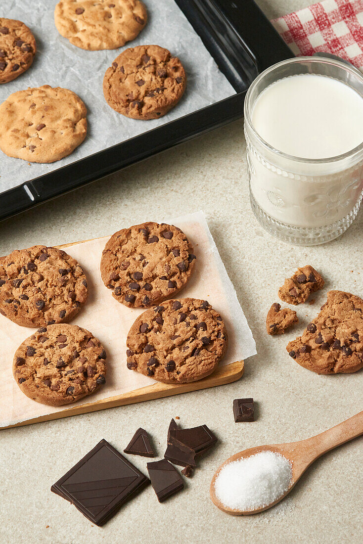 Fresh chocolate chip cookies on a wooden spatula beside a glass of milk, dark chocolate pieces, and a spoonful of sugar on a kitchen counter