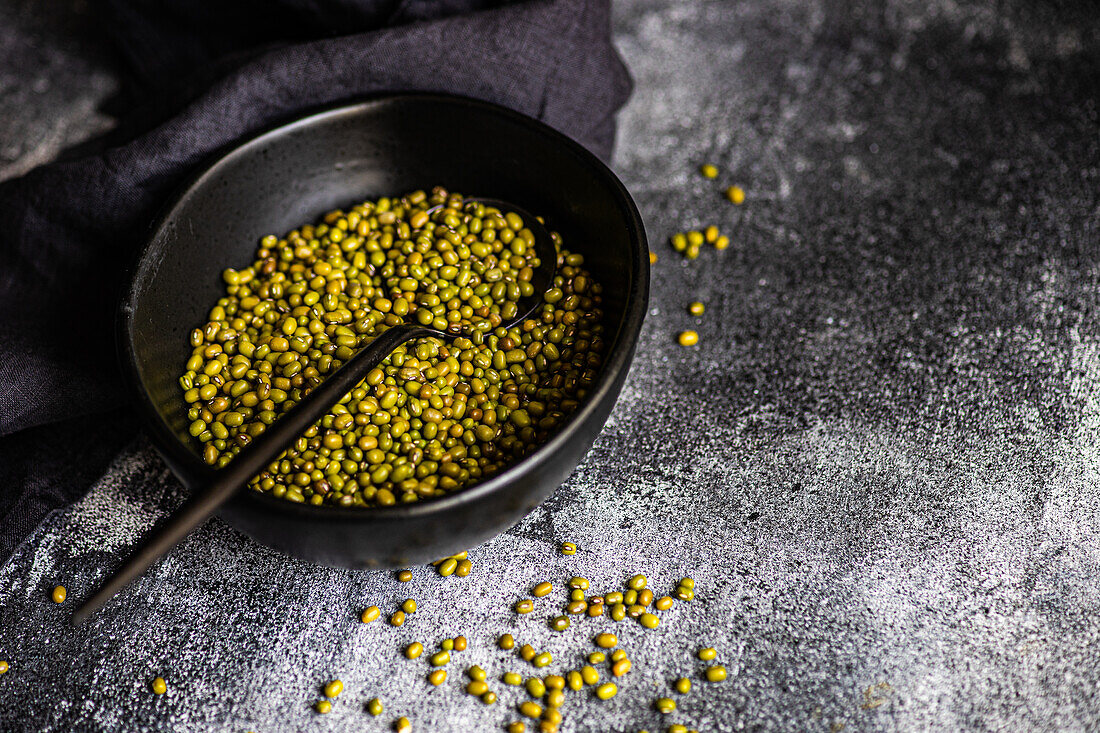 A rustic setting features raw organic mung beans in a textured black bowl, scattered loosely with a spoon, on a dark, grainy surface, highlighted by natural light