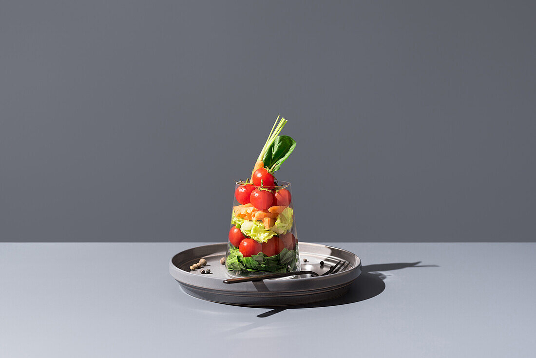 Creatively layered salad with tomatoes, carrots, and lettuce in a transparent glass, displayed elegantly on a gray background with stark shadows.