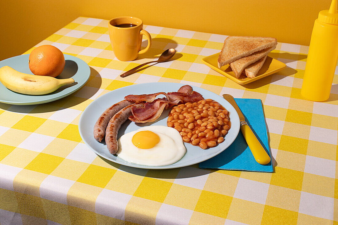 Yellow coloured Tablecloth with a delicious full English Breakfast, egg, beans, sausages and bacon, some toasted bread slices, fruit and a cup of coffee