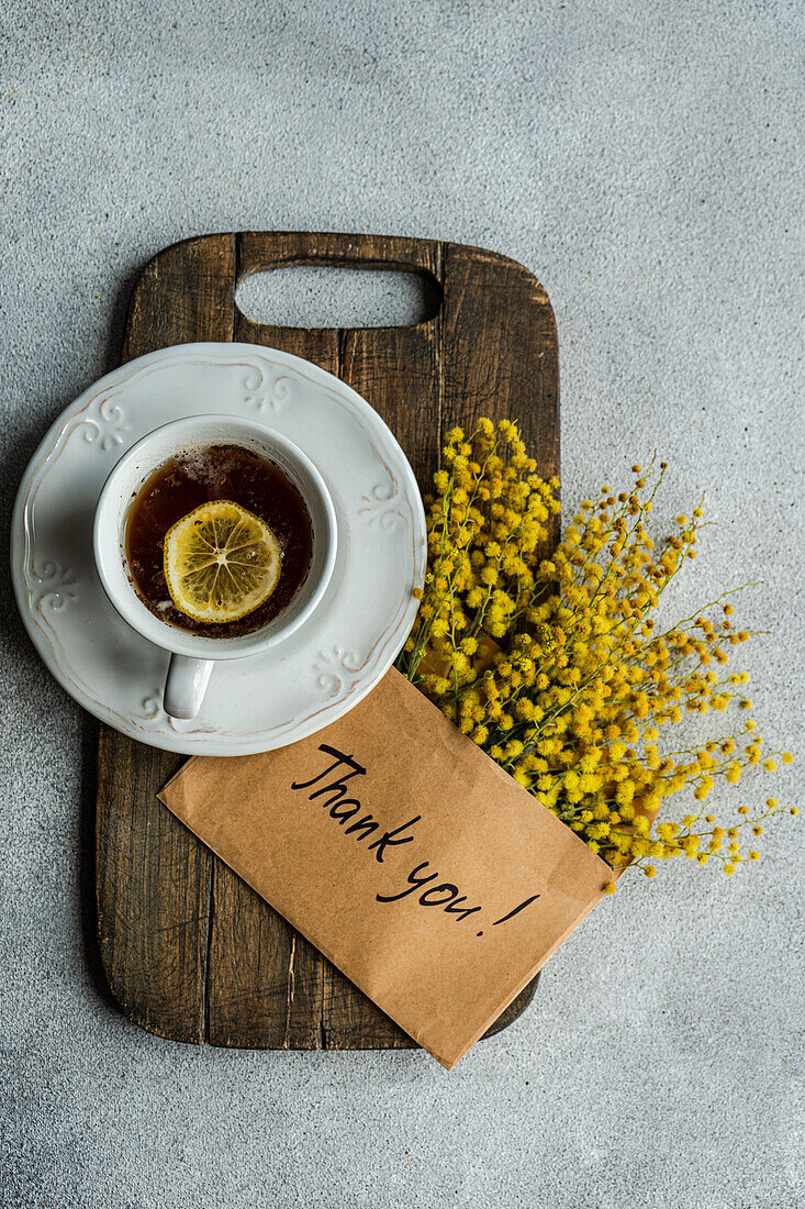 A top view of a classic tea cup with lemon, placed on a wooden board, accompanied by bright Mimosa flowers and a Thank You! card on a textured background