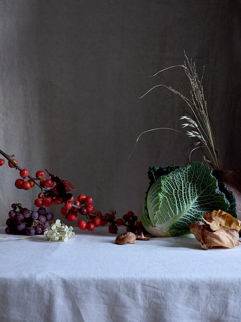 A tasteful arrangement featuring savoy cabbage, red grapes, and tiny red crab apples, artistically displayed on a draped white fabric with subtle lighting.