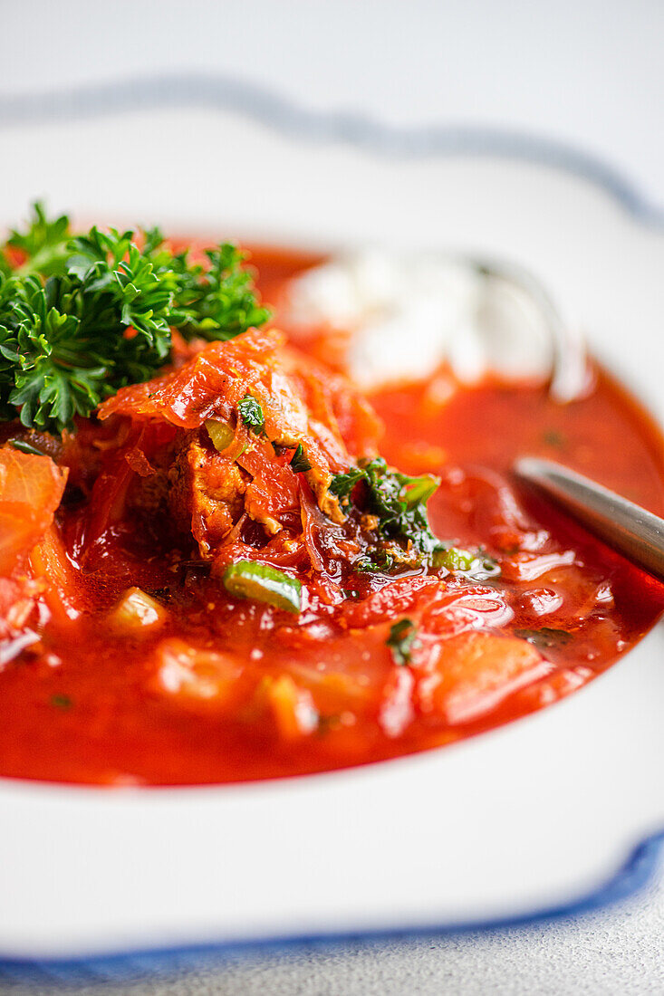 Nahaufnahme einer herzhaften Portion ukrainischen Borschtsch, einer traditionellen Rote-Bete-Suppe, die mit einem Klecks saurer Sahne und frischen Kräutern verfeinert und in einer weißen Schüssel mit einem Löffel an der Seite präsentiert wird