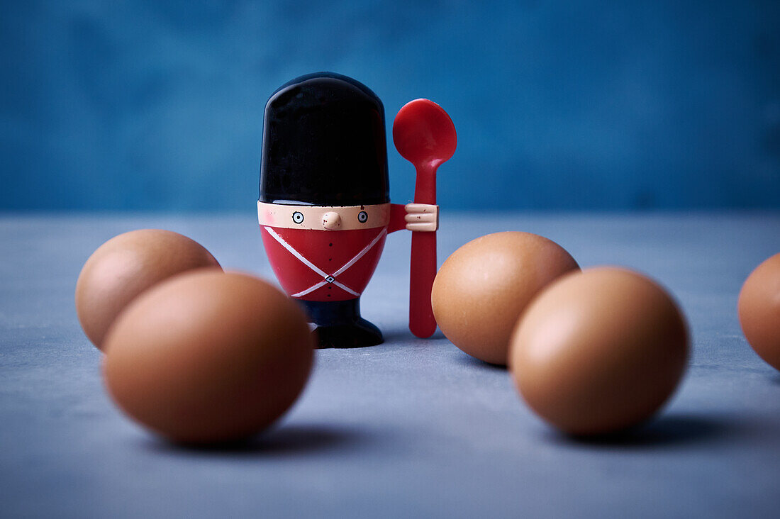 A whimsical soldier egg cup armed with a spoon stands guard among a formation of brown eggs on a soft blue background, creating a playful kitchen scene.