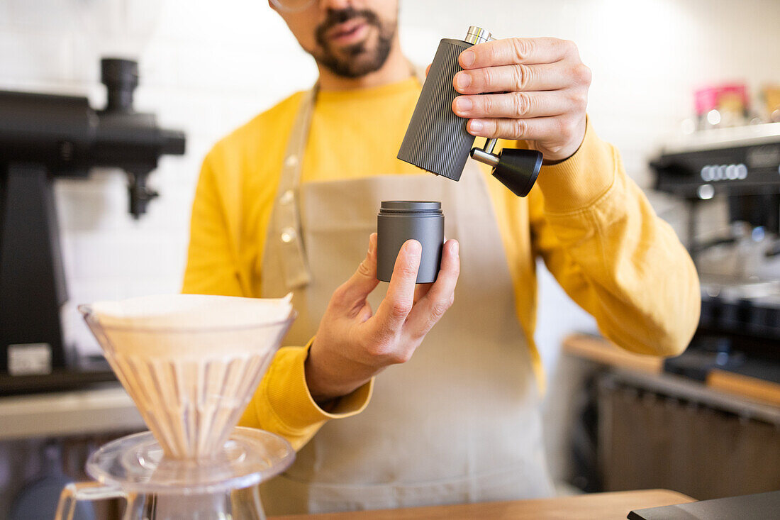 A barista in a yellow apron grinds fresh coffee beans using a manual grinder into a portable cup at a modern cafe.