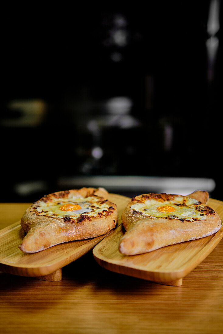 Close-up image capturing authentic Georgian Khachapuri, featuring a crispy crust, melted cheese, and a perfectly cooked egg, served on rustic wooden boards in a dimly lit setting