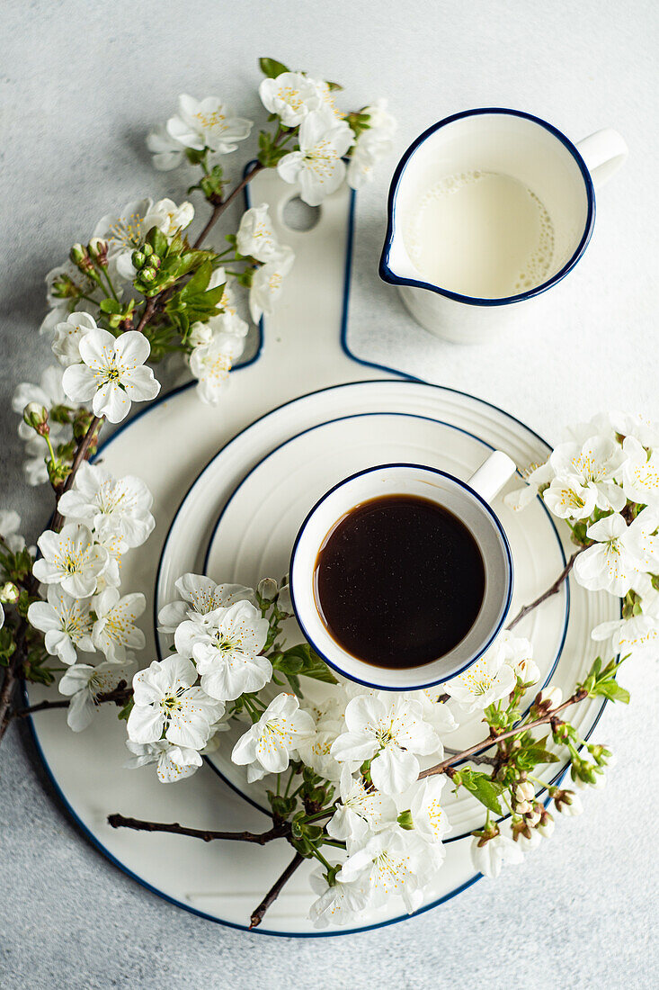 A cup of black coffee and fresh milk in a white enamel mug alongside cherry blossoms on a plate, capturing the essence of spring