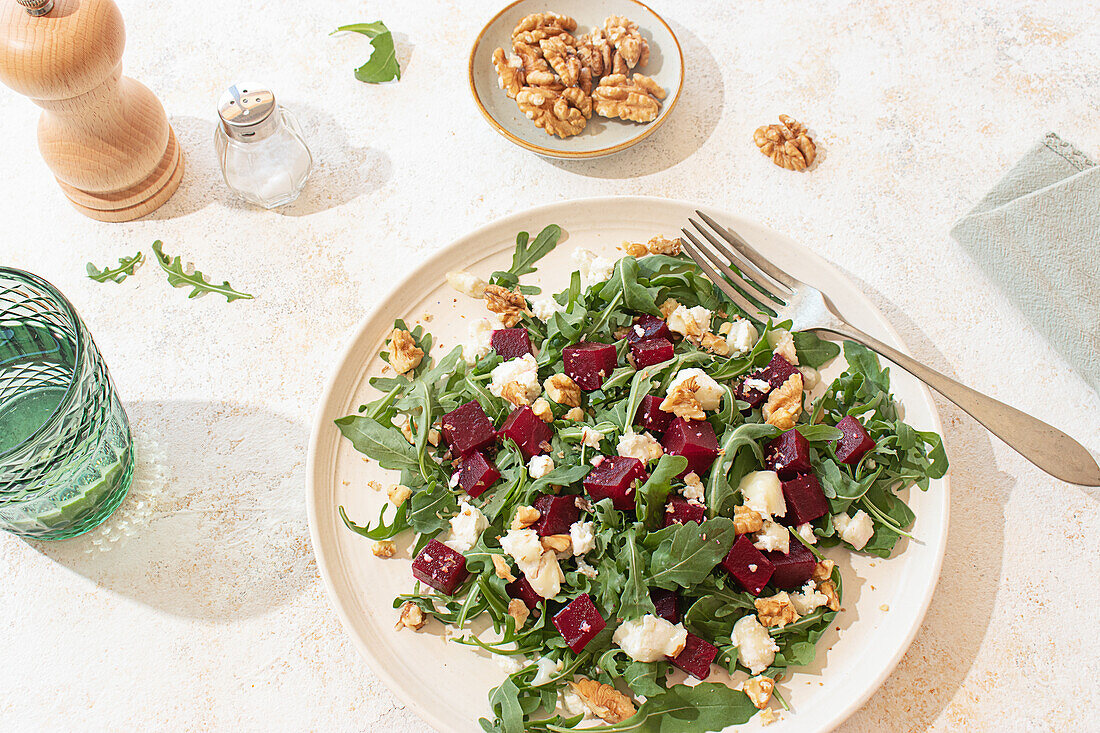 Healthy goat cheese, arugula and beetroot salad on a beige plate and styled with a pepper mill, a lime water glass on a rustic but vibrant atmosphere