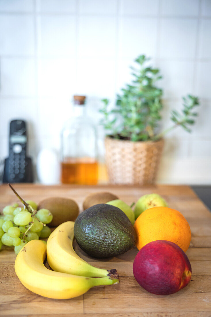 Eine Vielzahl von frischen Früchten, die auf einer hölzernen Küchentheke angeordnet sind. Diese Szene zeigt Bananen, eine Avocado, eine Mango, Kiwis, Weintrauben und eine Mango, aufgelockert durch einen Hintergrund mit natürlichem Licht und Küchenutensilien sowie einer Topfpflanze.