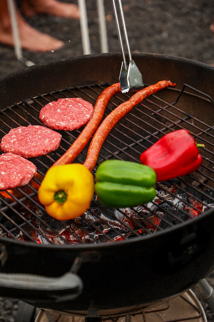 Cropped unrecognizable person grilling sausages and vegetables on a barbecue at an outdoor gathering. Background includes guests socializing, enjoying drinks and music.