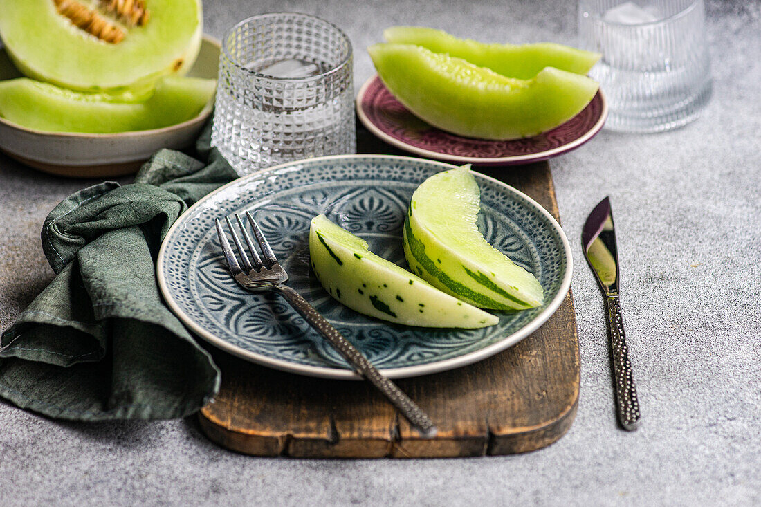 From above various organic mini melon slices artistically presented on an ornate blue patterned plate, coupled with a textured grey napkin and elegant cutlery; an ideal fresh and light lunch setup.