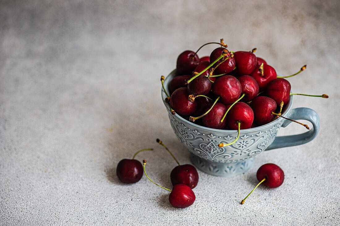 From above blue ornate ceramic bowl filled with fresh red cherries, situated on a textured grey surface. Several cherries are scattered around the bowl, enhancing the natural appeal.