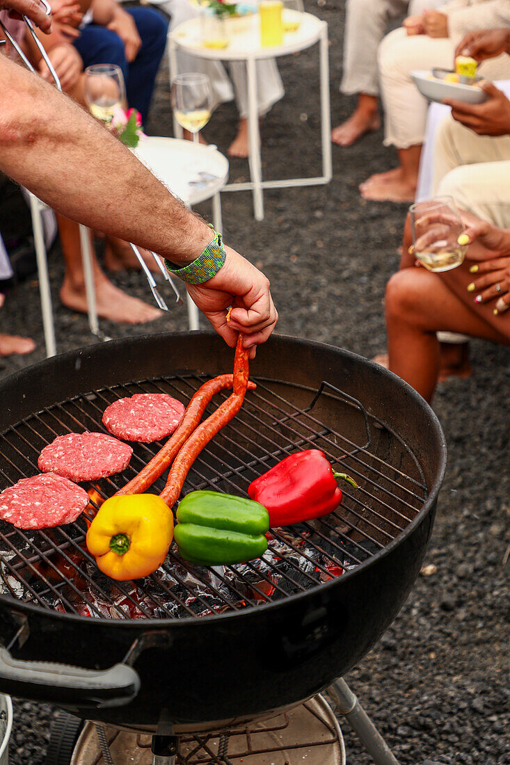 Cropped unrecognizable person grilling sausages and vegetables on a barbecue at an outdoor gathering. Background includes guests socializing, enjoying drinks and music.