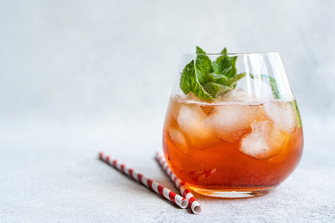 Gekühlter Cola-Cocktail, garniert mit frischen Minzblättern, serviert in einem Glas mit Eiswürfeln und zwei rot gestreiften Papierstrohhalmen, vor einem strukturierten grauen Hintergrund.