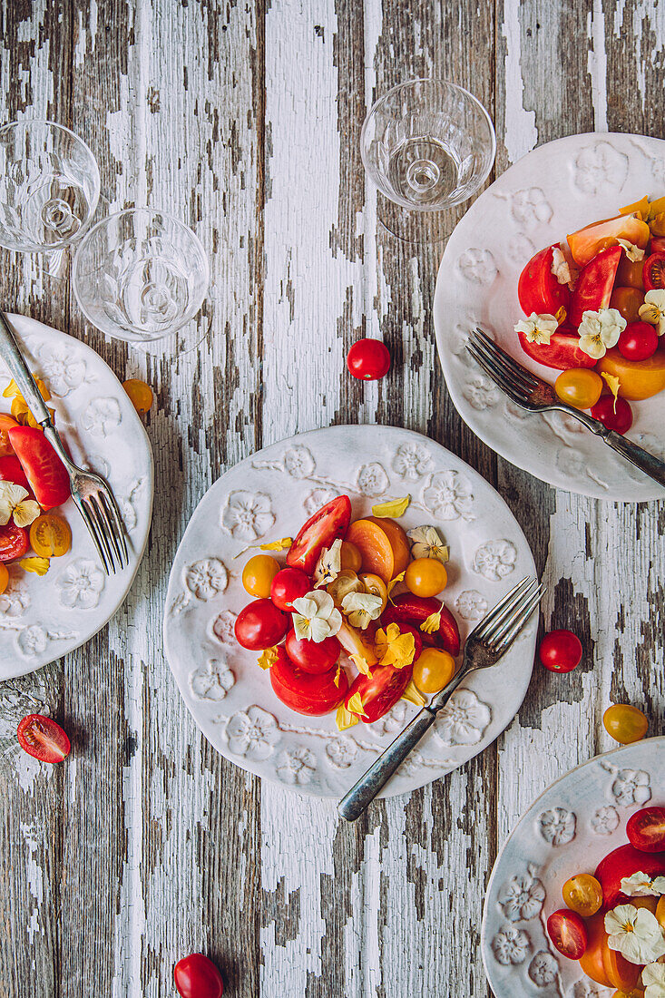 Elegant plates of colorful tomato salad with cherry tomatoes are ready to serve on a rustic, white-painted wooden table, accompanied by wine glasses