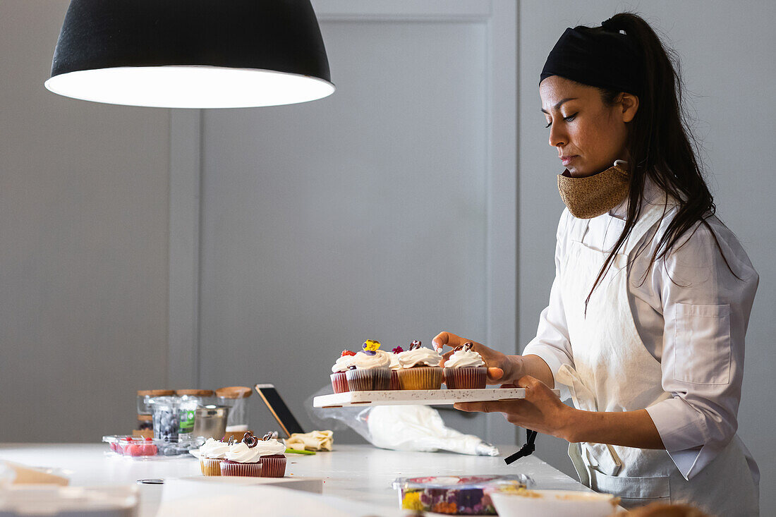 Side view of female baker in uniform standing at table in bakery with vegan cupcakes on tray