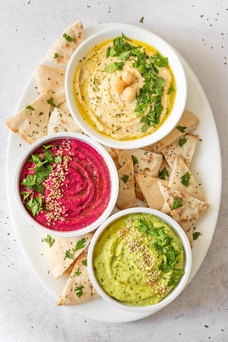 Draufsicht auf eine bunte Auswahl an klassischem, Rüben- und Avocado-Hummus, jeweils garniert und serviert mit Fladenbrot, als lebendiger, gesunder Dip für Snacks oder Vorspeisen.