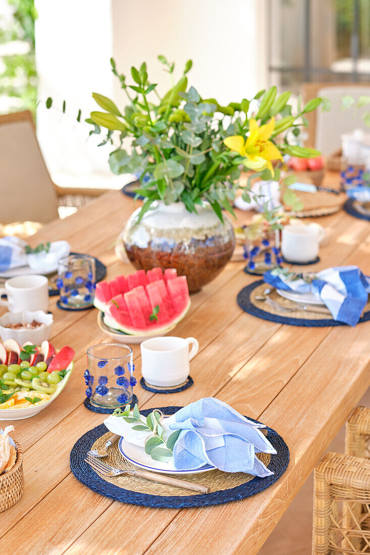 A vibrant and colorful breakfast set on a wooden terrace table, featuring a lush floral centerpiece and a selection of fresh fruits