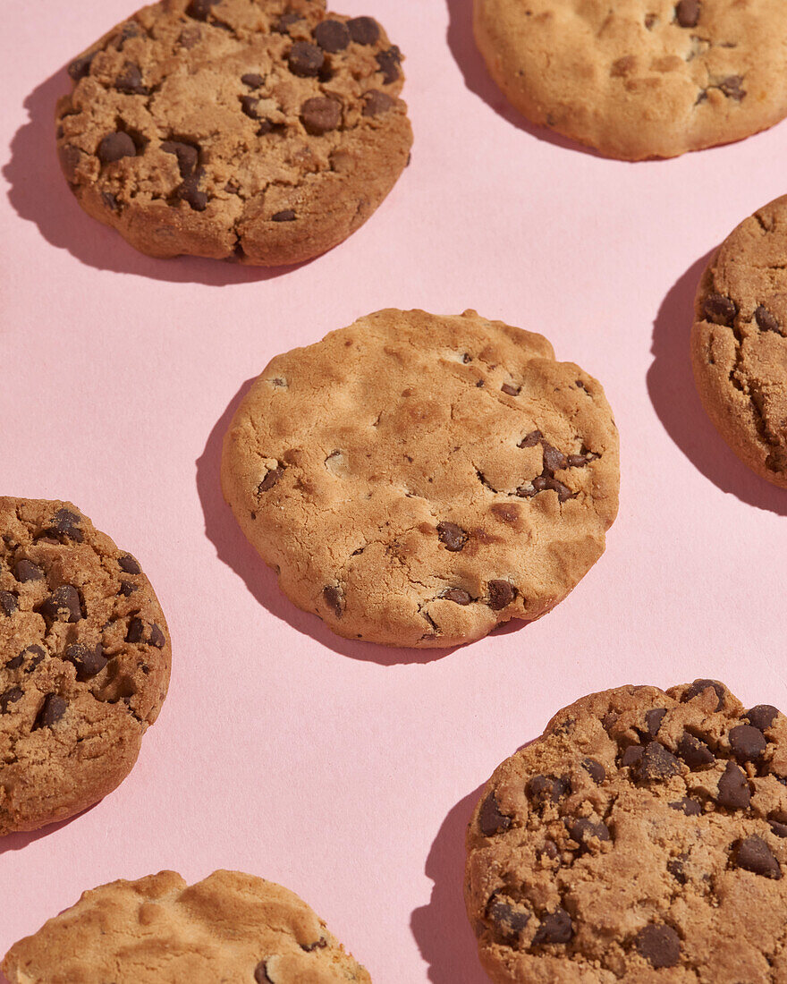 Scattered chocolate chip cookies on a pink surface, bathed in the warm glow of sunlight, creating inviting shadows